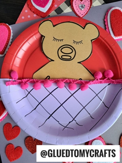 A sleeping teddy bear craft featuring a brown bear with closed eyes, tucked into a pink, basket-like paper plate with drawn lines and pink pom-poms, surrounded by red and pink felt heart decorations.