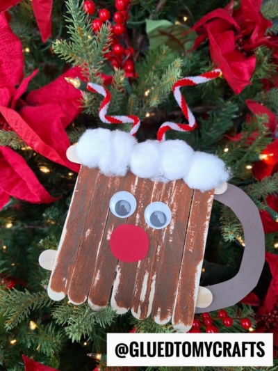 A handmade reindeer ornament, inspired by a cozy reindeer hot cocoa mug, is crafted from craft sticks, googly eyes, a red nose, cotton, and pipe cleaners. It hangs on a Christmas tree with red ribbons. @GLUEDTOMYCRAFTS is written at the bottom.