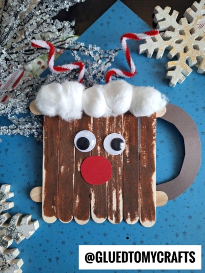 A handmade reindeer hot cocoa mug face craft made from popsicle sticks, with googly eyes, a red paper nose, cotton ball “hair,” and a curved paper handle, placed on a blue background with snowflake decorations.