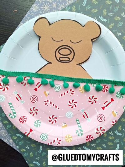A sleeping teddy bear craft featuring a bear snoozing under a pink, candy-patterned paper plate blanket. The brown head has a sweet drawn face, and green pom-poms trim the blanket’s edge. @GLUEDTOMYCRAFTS appears at the bottom.