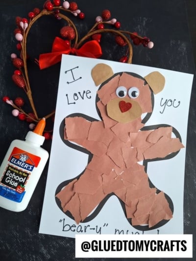 A handmade torn paper bear craft features brown paper, googly eyes, and a red heart nose on white paper that says "I love you bear-y much!" A glue bottle sits nearby, with a heart-shaped decoration in the background.