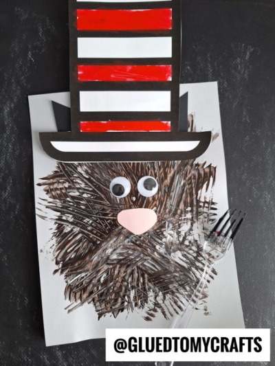A fork painted Cat in the Hat-inspired craft made from paper, featuring a red and white striped hat, googly eyes, a pink nose, and brown brushstrokes for fur. @GLUEDTOMYCRAFTS text label at the bottom.