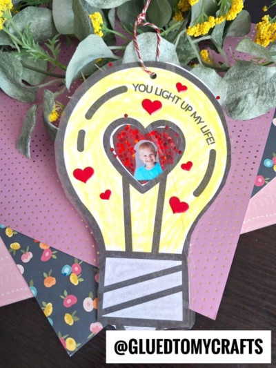 A handmade light bulb photo ornament card with the words “You light up my life!” Features a small photo of a child in a heart-shaped window, red heart decorations, and flowers and colored paper in the background. @GLUEDTOMYCRAFTS.