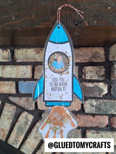 A handmade valentine's day rocket craft with a child's photo in the window, blue accents, glitter, and a handprint as flames, hanging by a red and white string. The text reads, Love you to the moon and back!.