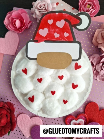 A paper plate Valentine gnome with a cotton ball beard, red paper hat, and small red heart shapes, surrounded by pink and red paper flowers and hearts for a festive touch.