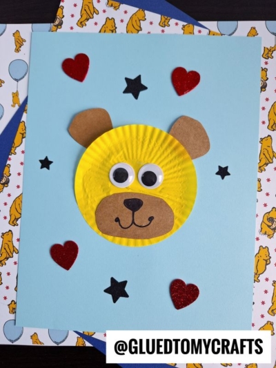 A handmade cupcake liner bear face card, featuring a yellow cupcake liner, googly eyes, and brown paper ears, nose, and mouth. Red hearts and black stars add charm to the blue background. @GLUEDTOMYCRAFTS is written at the bottom.