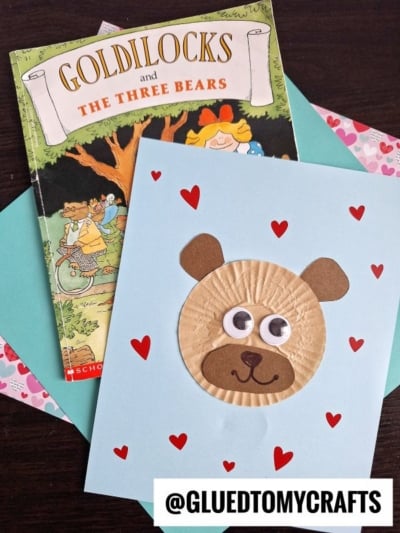 A handmade cupcake liner bear card with heart decorations and a Goldilocks and the Three Bears book in the background, displayed on a wooden table. @GLUEDTOMYCRAFTS is written at the bottom.