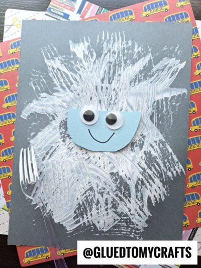 A dark sheet of paper features a fork painted yeti with googly eyes and a smile, alongside a white paint splatter made with a plastic fork, all atop school bus-themed paper. Text reads @GLUETOMYCRAFTS.