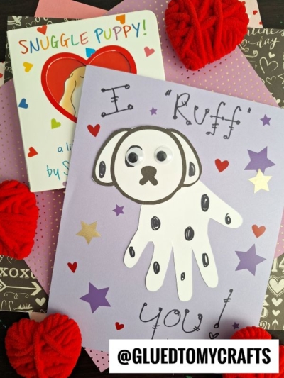 A handmade card with a white paper handprint dalmatian dog, googly eyes, and the text “I ‘Ruff’ You!” surrounded by colorful stars, sits on top of a heart-themed book and paper. Red yarn hearts are nearby. @GLUEDTOMYCRAFTS is at the bottom.