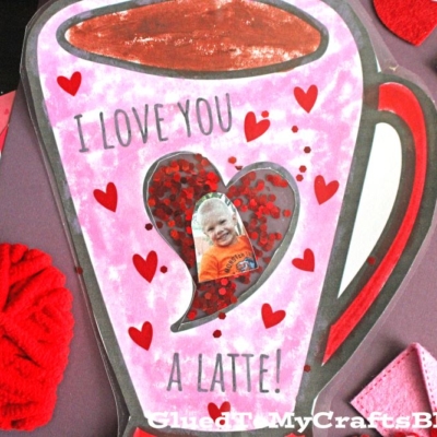 A handmade Valentine’s card shaped like a coffee cup with “LOVE YOU A LATTE PHOTO!” written on it, decorated with red hearts and featuring a child’s photo inside a heart-shaped window.