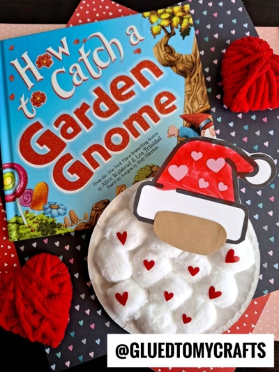 A colorful book titled How to Catch a Garden Gnome lies on heart-patterned paper, next to a paper plate valentine gnome craft with a cotton ball beard and red heart accents. Yarn hearts and a @GLUETOMYCRAFTS tag are visible.