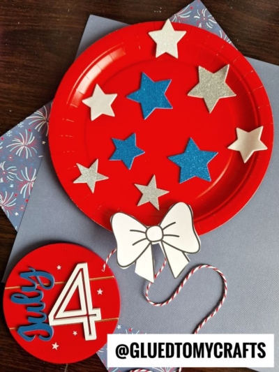 A red paper plate balloon decorated with silver and blue stars and a white bow, with July 4 written on a red circle nearby. The image features patriotic, Fourth of July-themed crafts on a blue background.