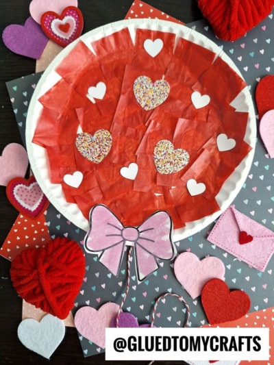 A paper plate balloon decorated with red tissue paper and heart shapes, featuring a pink paper bow at the bottom, surrounded by felt and paper hearts on a heart-patterned background. @GLUEDTOMYCRAFTS is written at the bottom.
