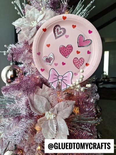 A pink Christmas tree adorned with pink flowers, white pearls, a disco ball ornament, and a paper plate balloon with hand-drawn hearts and a bow. Text at the bottom reads @GLUEDTOMYCRAFTS.