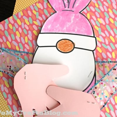 A handmade craft features a pink bunny gnome with paper arms, set against colorful patterned paper and ribbon.