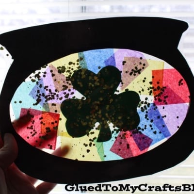 A hand holds up a pot-of-gold-shaped suncatcher made from the Free Black Cauldron Printable—Landing Page, decorated with colorful tissue paper, a green shamrock, and gold glitter, as sunlight shines through a window behind it.