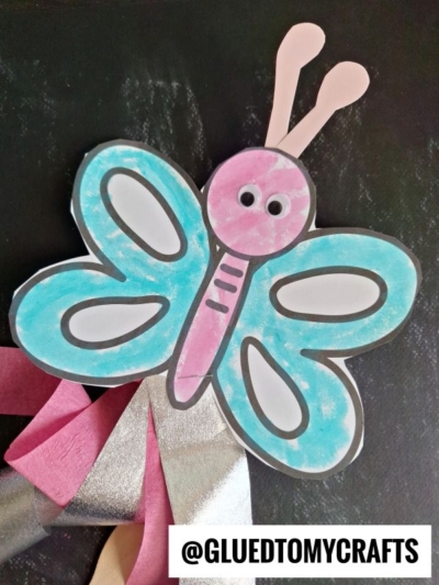 A colorful butterfly wand craft with blue wings, a pink body, googly eyes, and pink and silver streamers, displayed on a black background. The handle @GLUEDTOMYCRAFTS is visible at the bottom.