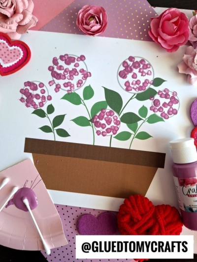 A Q-Tip Painted Hydrangea flowerpot craft with pink cotton swab flowers on white paper, surrounded by artificial blooms, felt hearts, craft paper, and a bottle of pink paint. @GLUEDTOMYCRAFTS is at the bottom.
