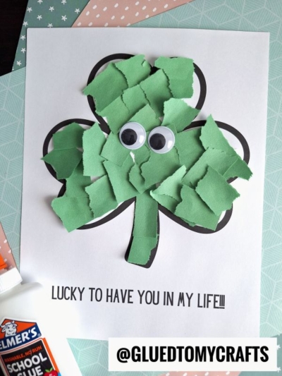 A torn paper shamrock outline is filled with green paper pieces and topped with two googly eyes. Text below reads, Lucky to have you in my life!!! An Elmers glue stick and patterned papers are nearby.