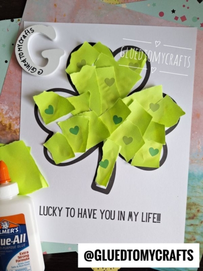 A torn paper shamrock is filled with green paper pieces and heart stickers. A bottle of glue and scrap paper sit nearby. Text on the page reads, LUCKY TO HAVE YOU IN MY LIFE!!.