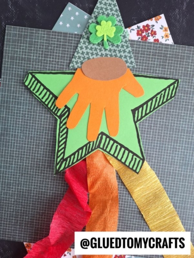 A star shaped leprechaun craft for St. Patrick’s Day, featuring a green star with striped borders, an orange handprint, and a brown oval. Topped with a green hat and clover, colorful streamers hang below. @GLUEDTOMYCRAFTS at the bottom.