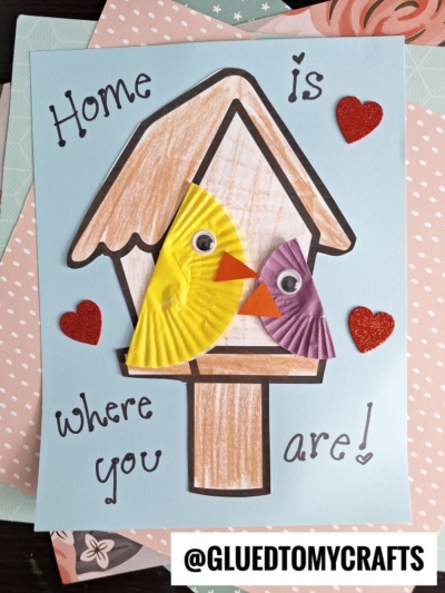 A handmade card with a birdhouse drawn in brown crayon, featuring a cupcake liner mama bird and chick (yellow and purple), googly eyes, red glitter hearts, and the text Home is where you are!.