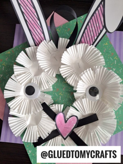A bunny face made from white cupcake liners, with large googly eyes, a pink paper heart nose, drawn bunny ears, and black ribbon whiskers—perfect alongside your next paper plate bunny wreath. Displayed on patterned paper. Text: @GLUEDTOMYCRAFTS.