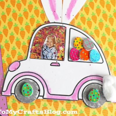 A handmade bunny car photo ornament shaped like a car with bunny ears, decorated with your child’s photo, colorful paper eggs, and cotton. Placed on carrot-patterned paper.