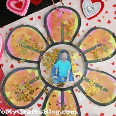 A flower photo ornament craft made from paper, with gold glitter on each petal and a child’s photo in the center.