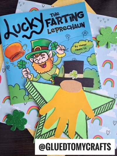 A children’s book titled Lucky the Farting Leprechaun lies on festive rainbow paper, with a star shaped leprechaun craft made of paper shapes and shamrock decorations beside it. A @GLUEDTOMYCRAFTS tag is at the bottom.