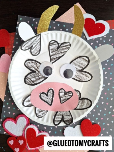 A charming paper plate cow craft decorated with black-outlined hearts, googly eyes, gold horns, and a pink snout, surrounded by red and pink paper hearts. @GluedToMyCrafts is written at the bottom.