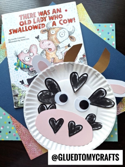 A children's book titled There Was an Old Lady Who Swallowed a Cow! is shown with a playful paper plate cow craft made from colored paper and heart shapes, placed on vibrant sheets. @GLUEDTOMYCRAFTS is labeled at the bottom.