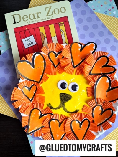 A childrens book titled Dear Zoo sits on patterned paper next to a cupcake liner lion craft made with orange cupcake liners, heart-shaped mane, googly eyes, and a yellow face. @GLUEDTOMYCRAFTS text is at the bottom.
