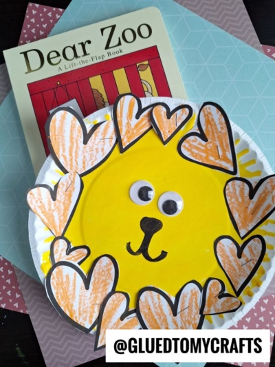 A “Dear Zoo” book lies on patterned paper beside a cupcake liner lion craft made from a yellow paper plate with a drawn-on face, googly eyes, and an orange heart-shaped mane. @GLUEDTOMYCRAFTS appears at the bottom.