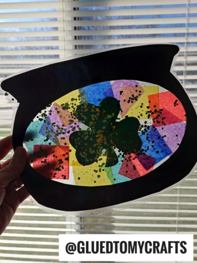 A hand holds up a leprechaun cauldron paper pot of gold craft with a shamrock cutout, filled with colorful tissue paper and glitter, against window blinds. Text at bottom reads @GLUEDTOMYCRAFTS.