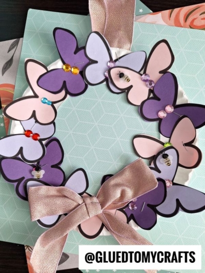 A handmade butterfly wreath features layered paper butterflies in purple and pink shades, colorful sequins, and a large pink ribbon bow, all resting on a pastel green patterned background. @GLUEDTOMYCRAFTS is written at the bottom.