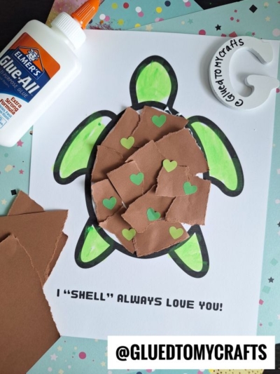 A torn paper turtle with a brown, textured shell and green heart stickers sits on a white sheet that reads I SHELL ALWAYS LOVE YOU! Elmers glue and craft supplies are scattered nearby.