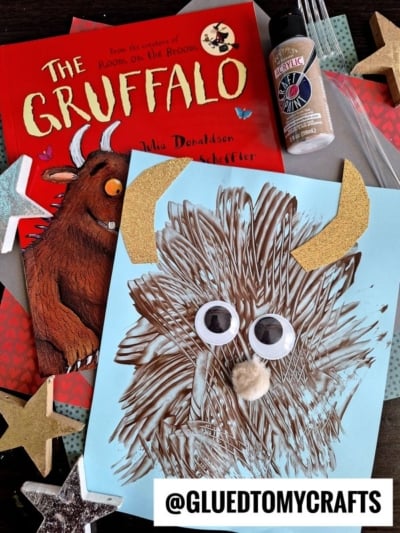 A fork painted Gruffalo book craft with googly eyes and gold horns on blue paper, made with acrylic paint, glitter, craft stars, and glue. Handle @GLUEDTOMYCRAFTS at the bottom.