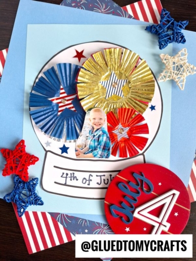 A child’s photo is inside a patriotic snow globe craft, decorated with red, white, blue, and gold cupcake liners shaped like fireworks. String stars and July 4 embellishments surround this festive creation.