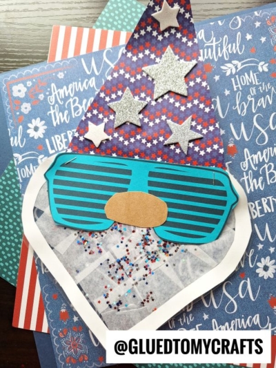 A handmade paper summer gnome suncatcher with blue striped sunglasses, a brown nose, a hat adorned with silver stars, and a beard dusted in red, white, and blue glitter on patriotic-themed paper. @GLUEDTOMYCRAFTS is at the bottom.