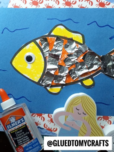 A torn paper fish with foil scales and a googly eye is glued to a blue background. Nearby are Elmer’s glue, a mermaid cutout, and a label reading @GLUEDTOMYCRAFTS. Red crab patterns border the image.