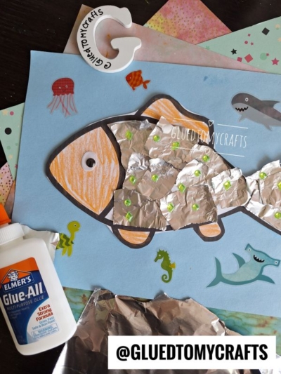 A children’s craft project featuring a torn paper fish made from paper and aluminum foil on blue paper, with sea creatures drawn around it. Elmer’s glue, foil, and craft supplies are scattered nearby.