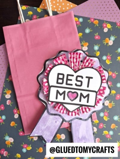 A handmade best mom badge ribbon in pink and purple rests on a pink gift bag, placed over floral and polka dot patterned papers. The text @gluedtomycrafts appears at the bottom.