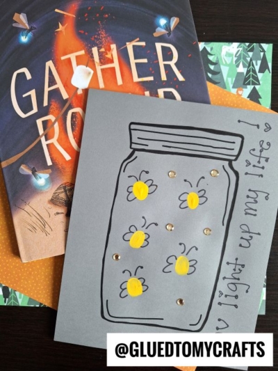 A handmade card shows a jar on gray paper, filled with fingerprint firefly art in yellow. Beside it is a Gather Round book and patterned paper. The card reads, You light up my life! with @GLUEDTOMYCRAFTS at the bottom.