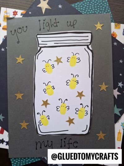 A craft project featuring a paper jar filled with yellow fingerprint fireflies, drawn wings and faces, and gold star stickers. The text says You light up my life. The handle @GLUEDTOMYCRAFTS is at the bottom.