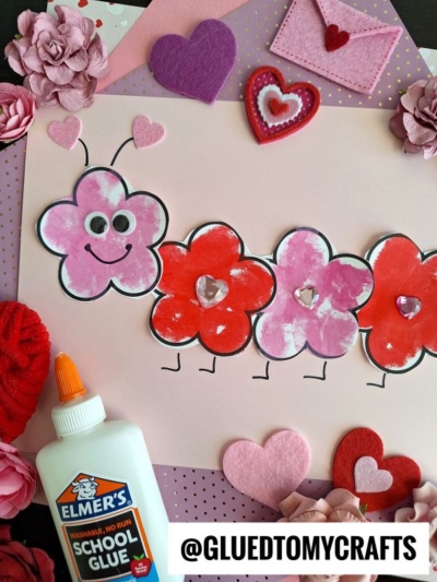 A cute flower caterpillar made from pink and red heart-shaped cutouts sits on a purple and pink background with heart and flower decorations. A bottle of Elmers school glue is in the corner, with “@GLUEDTOMYCRAFTS” at the bottom.