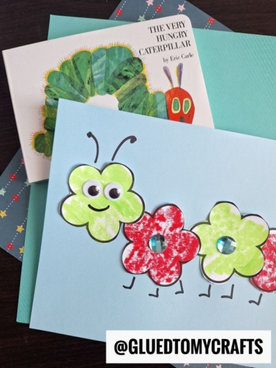 A handmade flower caterpillar card with three flower-shaped, green and red body segments, googly eyes, and a smile sits on top of The Very Hungry Caterpillar book. The handle @GLUEDTOMYCRAFTS appears at the bottom.