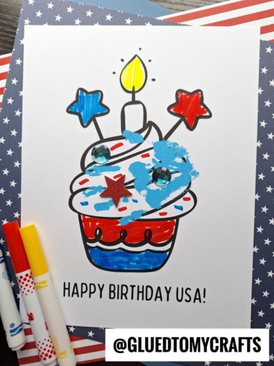 A handprint patriotic cupcake coloring page with HAPPY BIRTHDAY USA! text, decorated in red, blue, and white with star toppings, blue paint, and gems. Markers and a stars-and-stripes paper are nearby. @GLUEDTOMYCRAFTS is labeled.