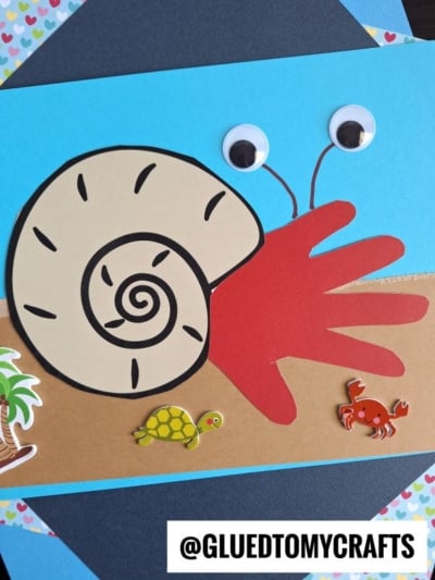 A colorful handprint hermit crab craft made with a red paper handprint, spiral seashell, googly eyes, and stickers of a turtle, crab, and palm tree on a blue paper background.