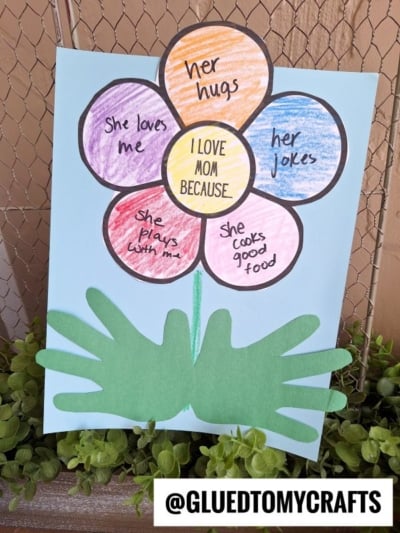A handmade Mother’s Day craft features a flower with colorful petals, each labeled "I love my mom because" with heartfelt reasons, on a blue background and green paper handprint leaves. The Instagram handle @GLUEDTOMYCRAFTS is at the bottom.